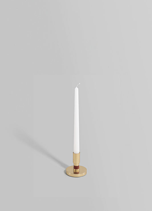 A tall, slender candle with a white wax body and a slightly curled wick, placed in a cylindrical holder combining gold and reddish-brown materials. The candle and holder are set against a plain light gray background.