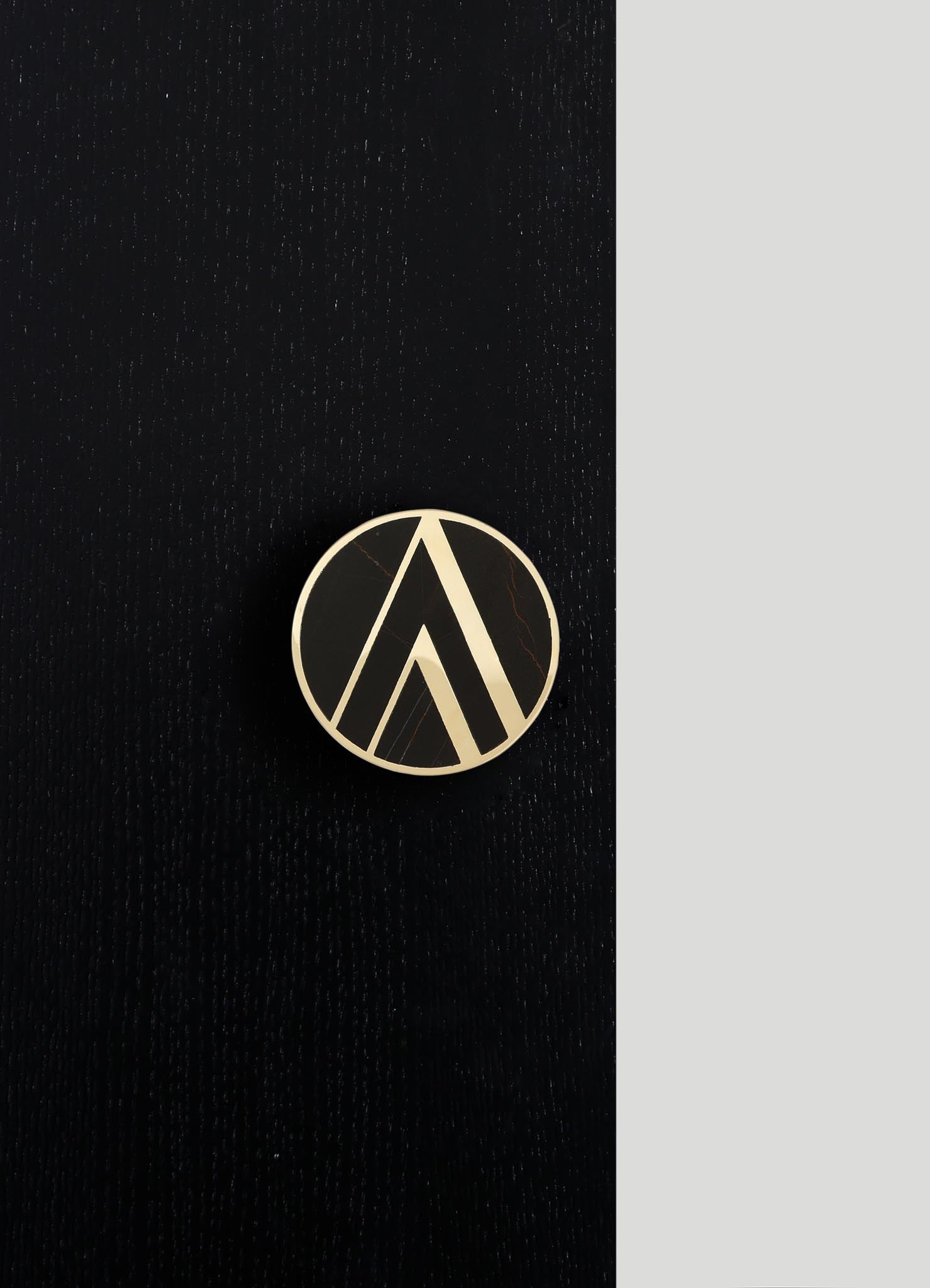A round brooch with geometric black and beige patterns, featuring angular lines and a polished finish, placed on a textured black surface with a contrasting gray background.