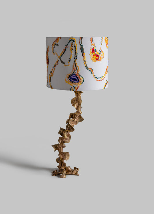 Table lamp with a sculptural brass base resembling abstract shapes and a fabric lampshade featuring embroidered multicolored abstract motifs on a white background.