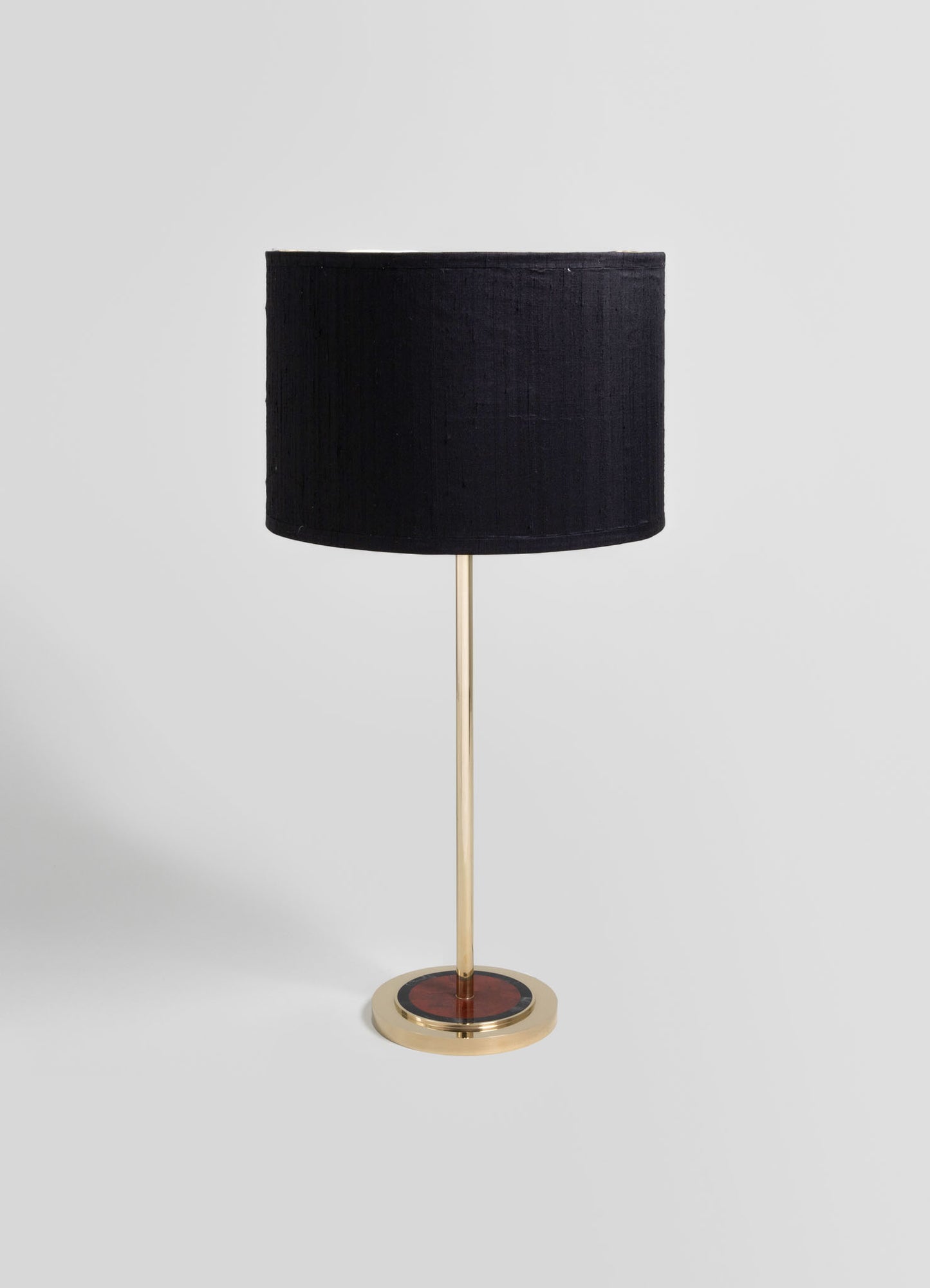 A table lamp with a black fabric drum shade and a slender brass stand, featuring a circular base with a red and black mosaic pattern in a contemporary style.