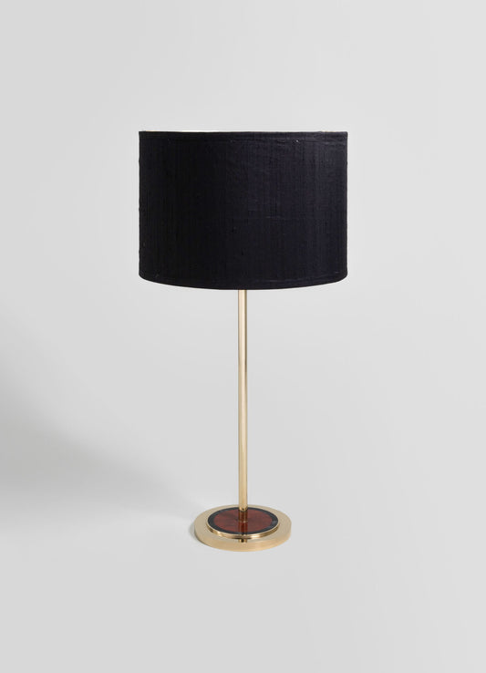 A table lamp with a black fabric drum shade and a slender brass stand, featuring a circular base with a red and black mosaic pattern in a contemporary style.