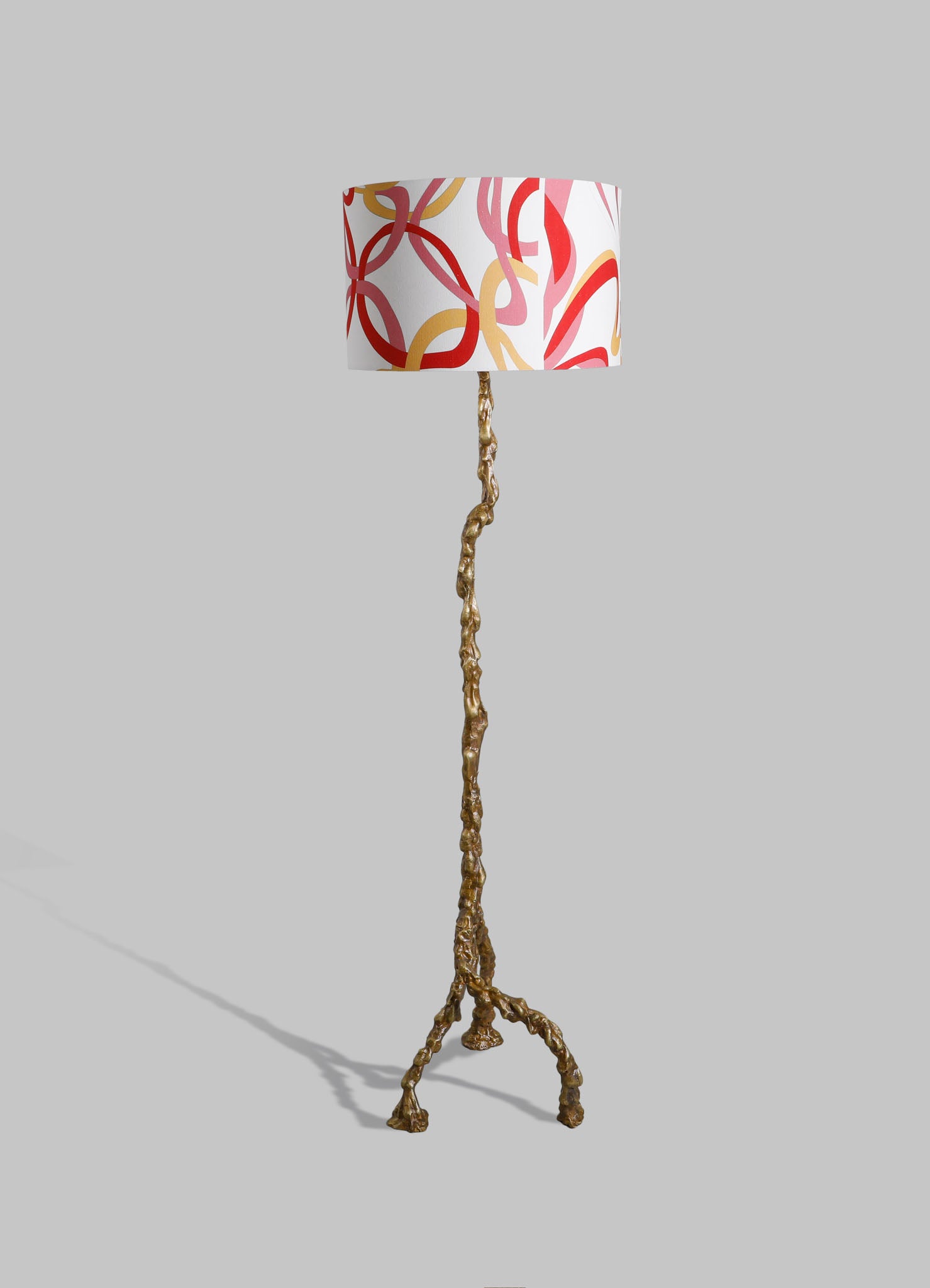Decorative table lamp with a textured, gold-colored, branch-like base and a cylindrical fabric shade featuring a bold red, pink, and beige abstract pattern on a white background.