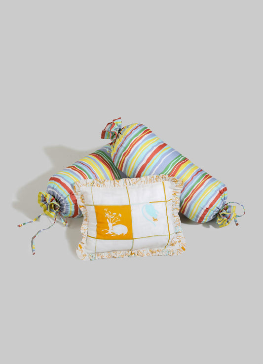 A set of baby nest and pillow with colorful striped fabric in shades of red, yellow, green, blue, and white, featuring tied ends and ruffled edges. The pillow has embroidered motifs of a rabbit and a bird, with a white background and yellow accents, all made of soft fabric suitable for nursery decor.