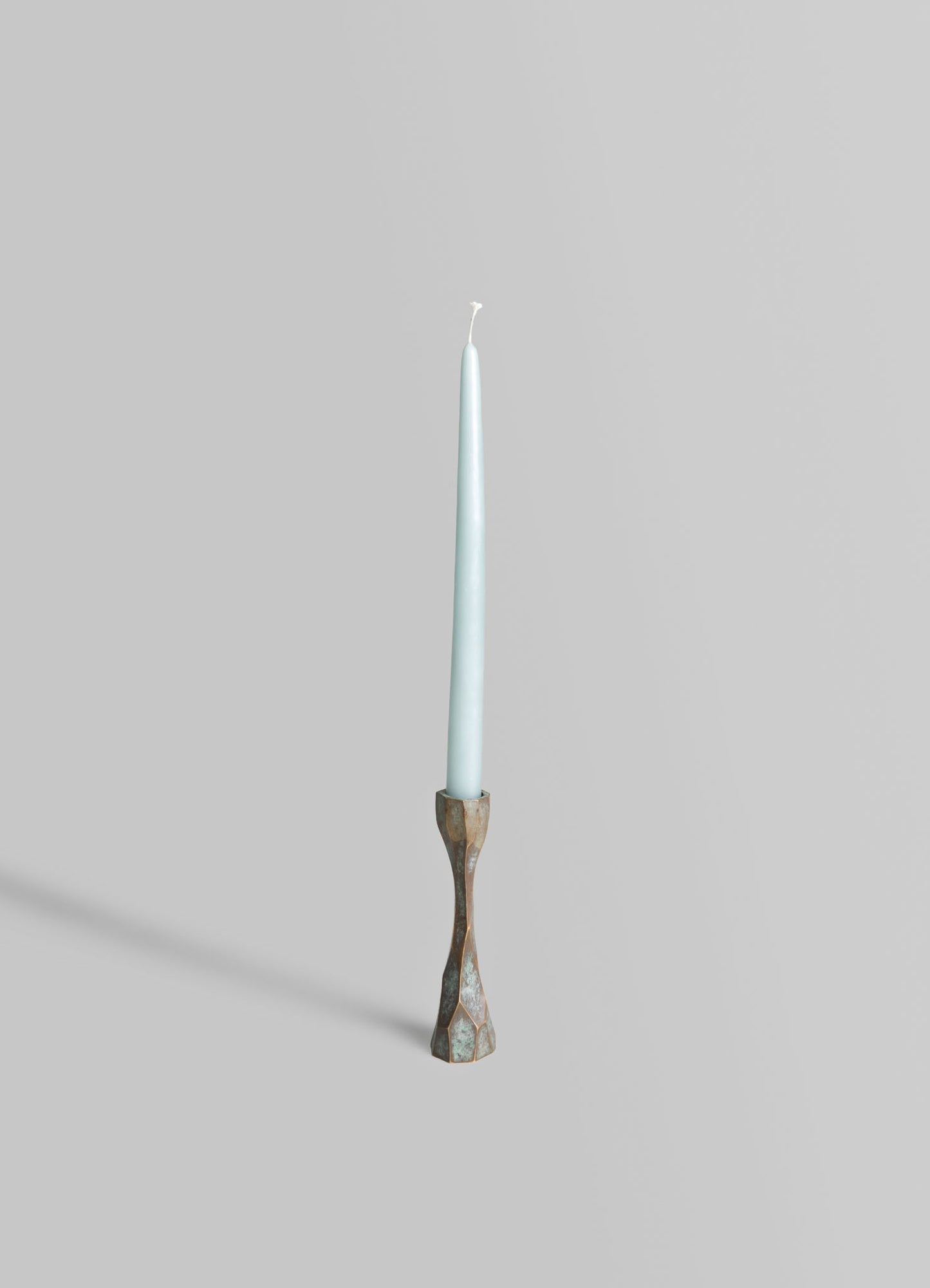 A tall, slender candle with a light blue finish placed in a sculptural brass candlestick holder with a faceted, geometric design. The scene has a minimalistic and contemporary aesthetic against a plain, light gray background.