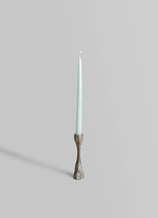 A tall, slender candle with a light blue finish placed in a sculptural brass candlestick holder with a faceted, geometric design. The scene has a minimalistic and contemporary aesthetic against a plain, light gray background.