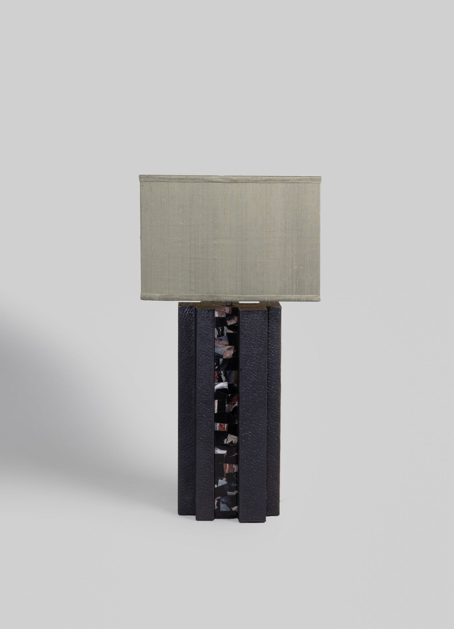 A table lamp featuring a rectangular fabric shade in neutral beige tones, mounted on a black base with a textured surface and a central decorative strip of glossy, irregularly shaped mosaic tiles.