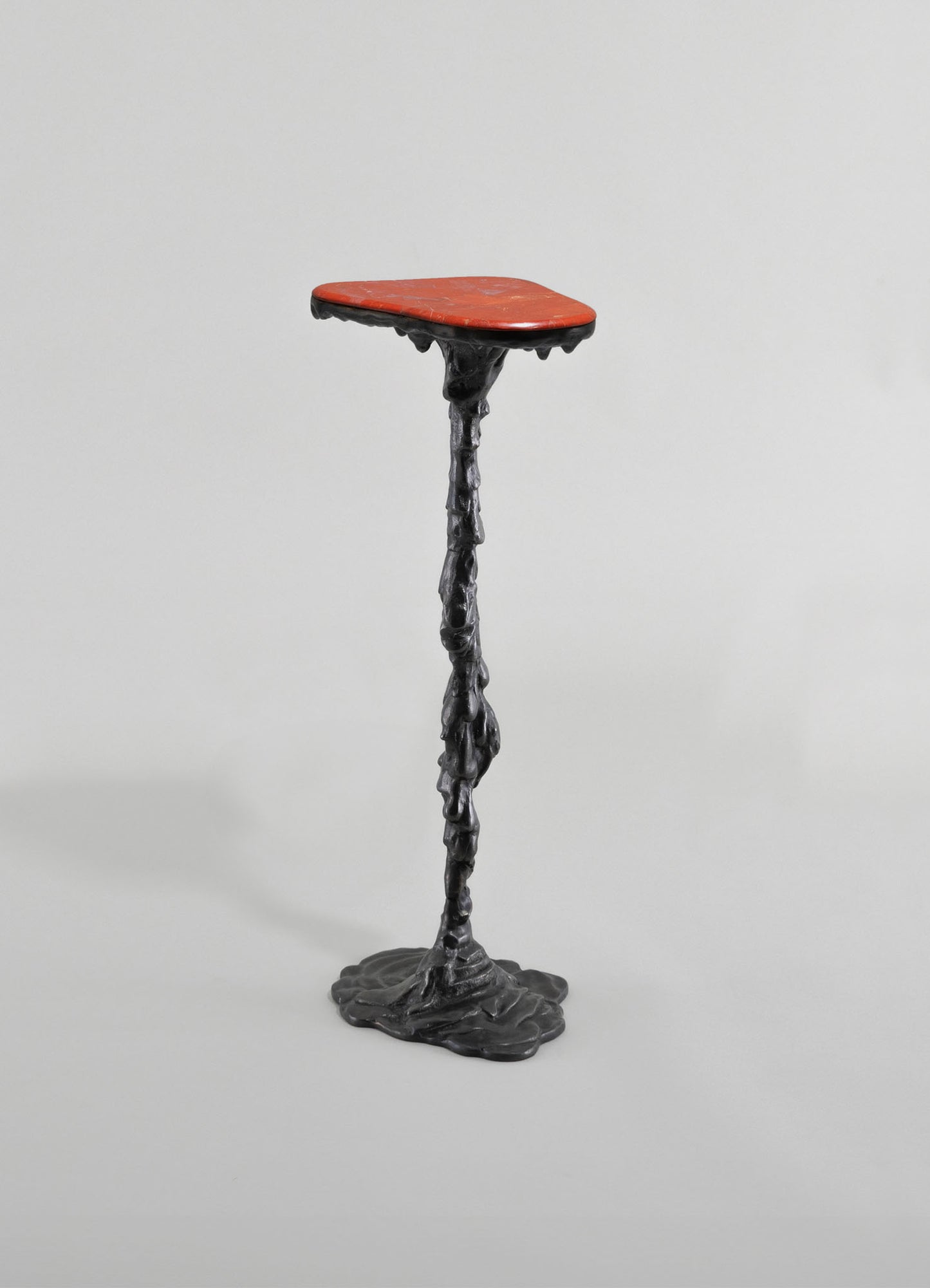 A sculptural side table with a textured black metal base resembling melted wax and an irregular, organic shape, topped with a smooth, oval-shaped red stone surface.
