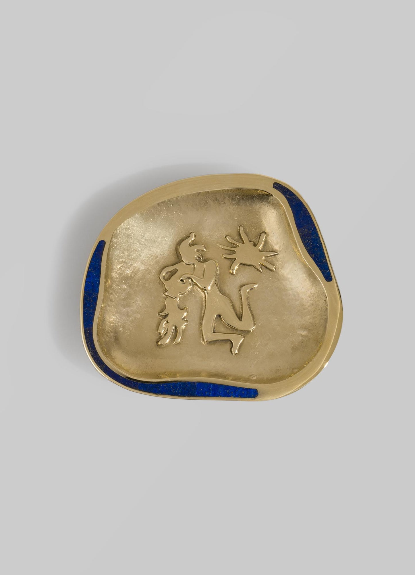 A decorative brass dish with an irregular shape and a textured surface features a raised abstract line drawing of a face. The dish has blue enamel accents along the edges, enhancing its artistic and handcrafted appearance.