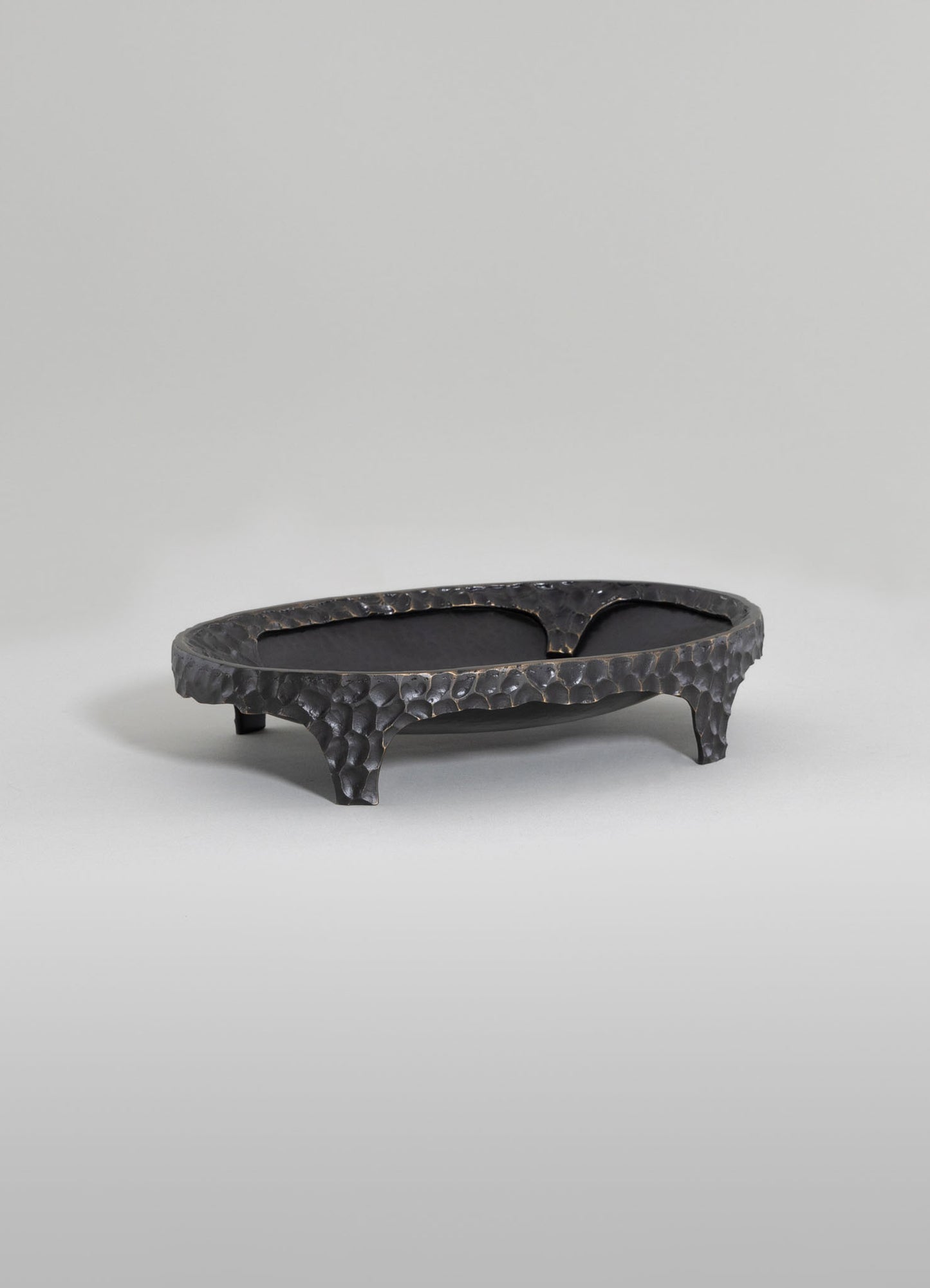 A handcrafted black metal tray with a hammered texture, featuring an oval shape and three curved legs, set on a plain light background.