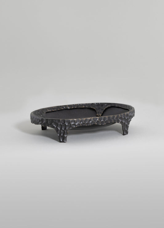 A handcrafted black metal tray with a hammered texture, featuring an oval shape and three curved legs, set on a plain light background.