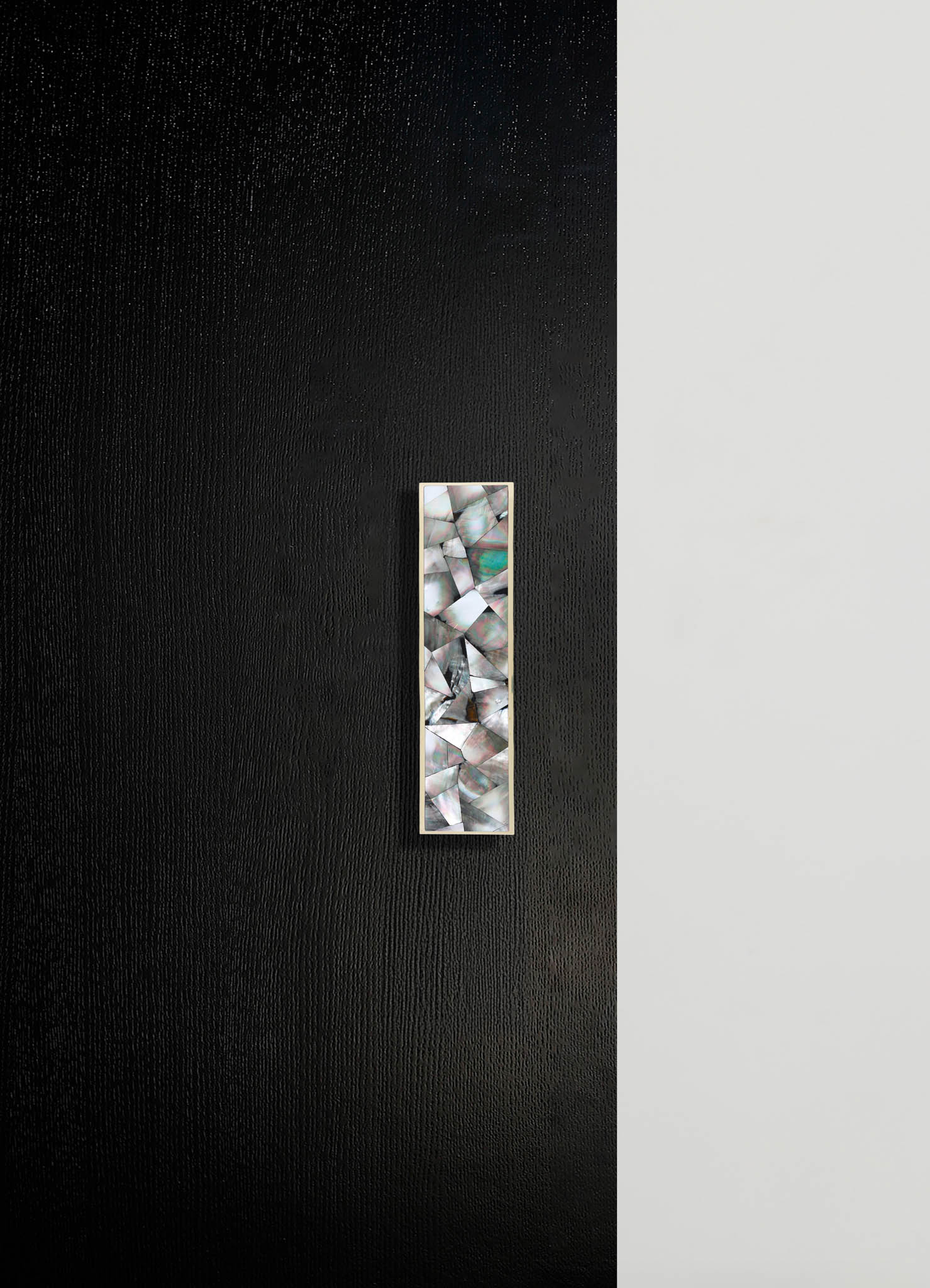 A vertical decorative panel with an iridescent, geometric mosaic pattern framed in silver, mounted on a two-tone wall with textured black on the left and smooth light gray on the right. The mosaic features irregular, shimmering shapes resembling shell or mother-of-pearl.
