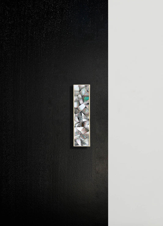 A vertical decorative panel with an iridescent, geometric mosaic pattern framed in silver, mounted on a two-tone wall with textured black on the left and smooth light gray on the right. The mosaic features irregular, shimmering shapes resembling shell or mother-of-pearl.