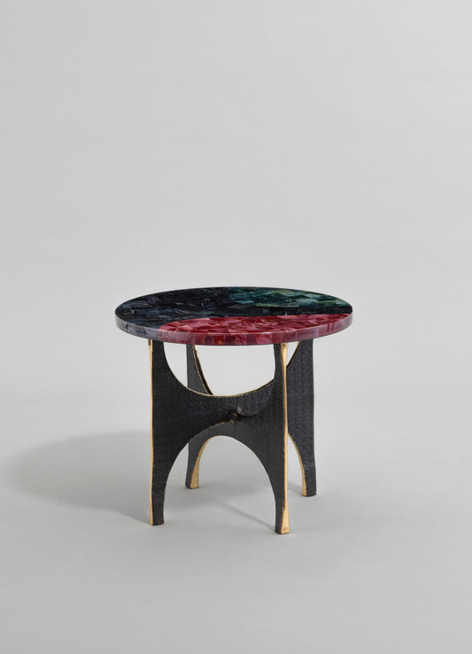 A small accent table with a round top made of multicolored mosaic glass in shades of black, green, and red, supported by black wooden legs with gold accents, featuring an arched design and a rustic, handcrafted style.