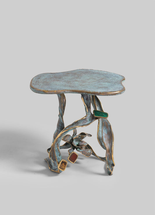 A sculptural side table crafted from metal with a distressed, oxidized finish and gold accents, featuring an organic, flowing form with a flat, irregularly shaped top and intertwined, leaf-like legs. The table includes embedded mineral stones in green, red, and brown tones, adding a natural motif.
