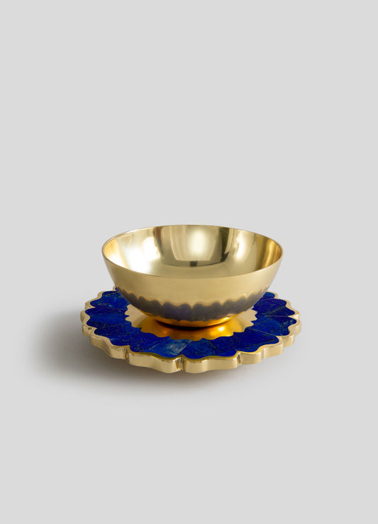 A gold-colored metal bowl with a smooth, reflective surface, placed on a decorative blue and gold ceramic coaster with a scalloped edge.