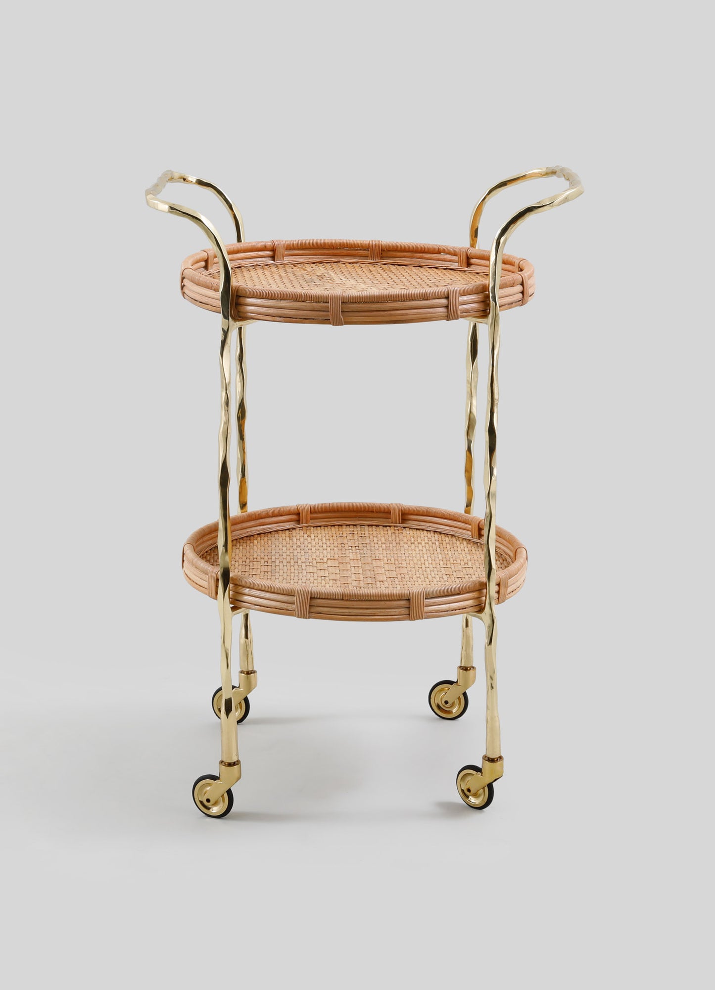 The VIYA Assam Brass & Cane Trolley features two round woven cane shelves, forged brass rods and legs, and four small wheels, all set against a plain light gray background.