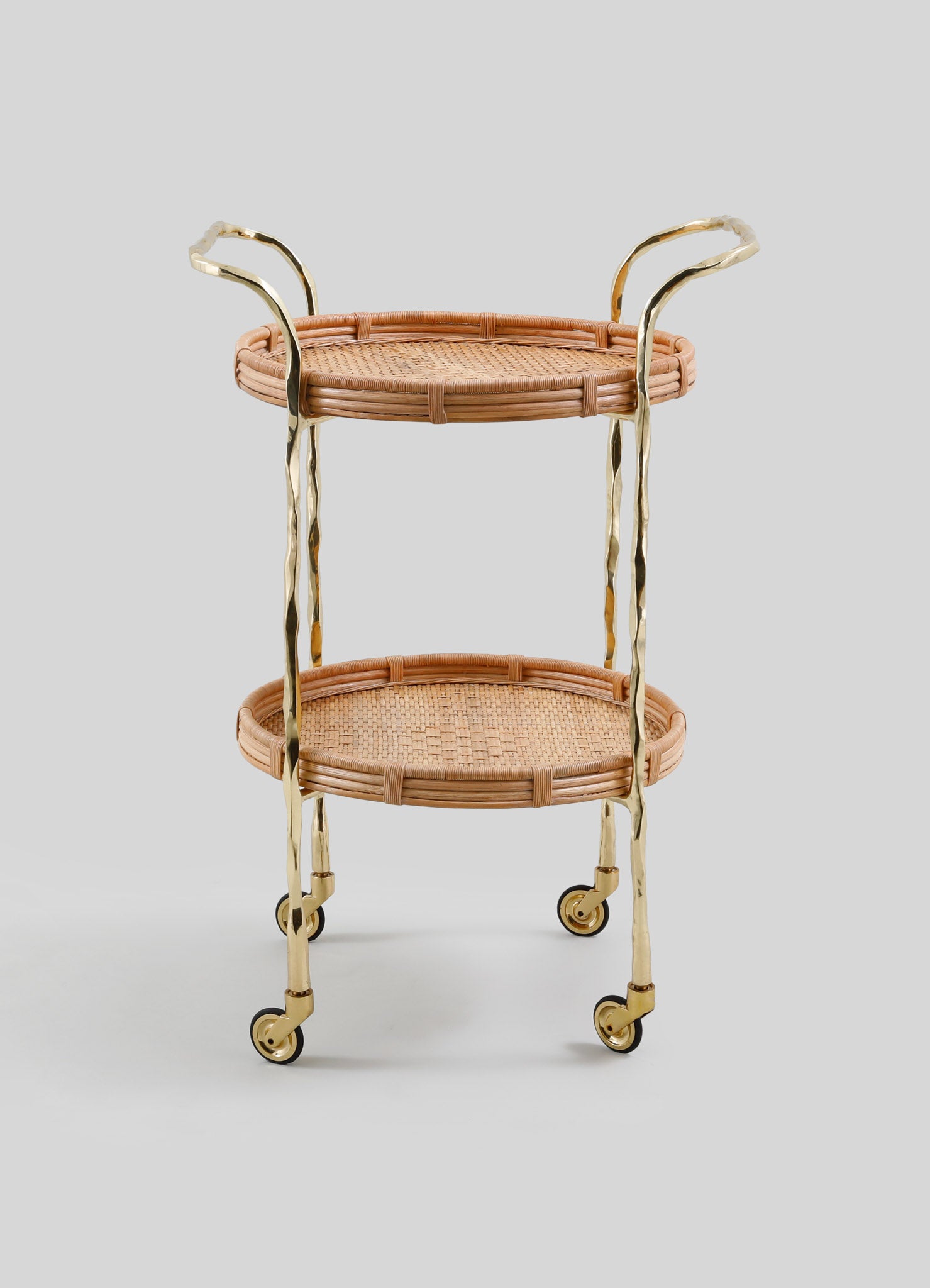 The VIYA Assam Brass & Cane Trolley features two round woven cane shelves, forged brass rods and legs, and four small wheels, all set against a plain light gray background.