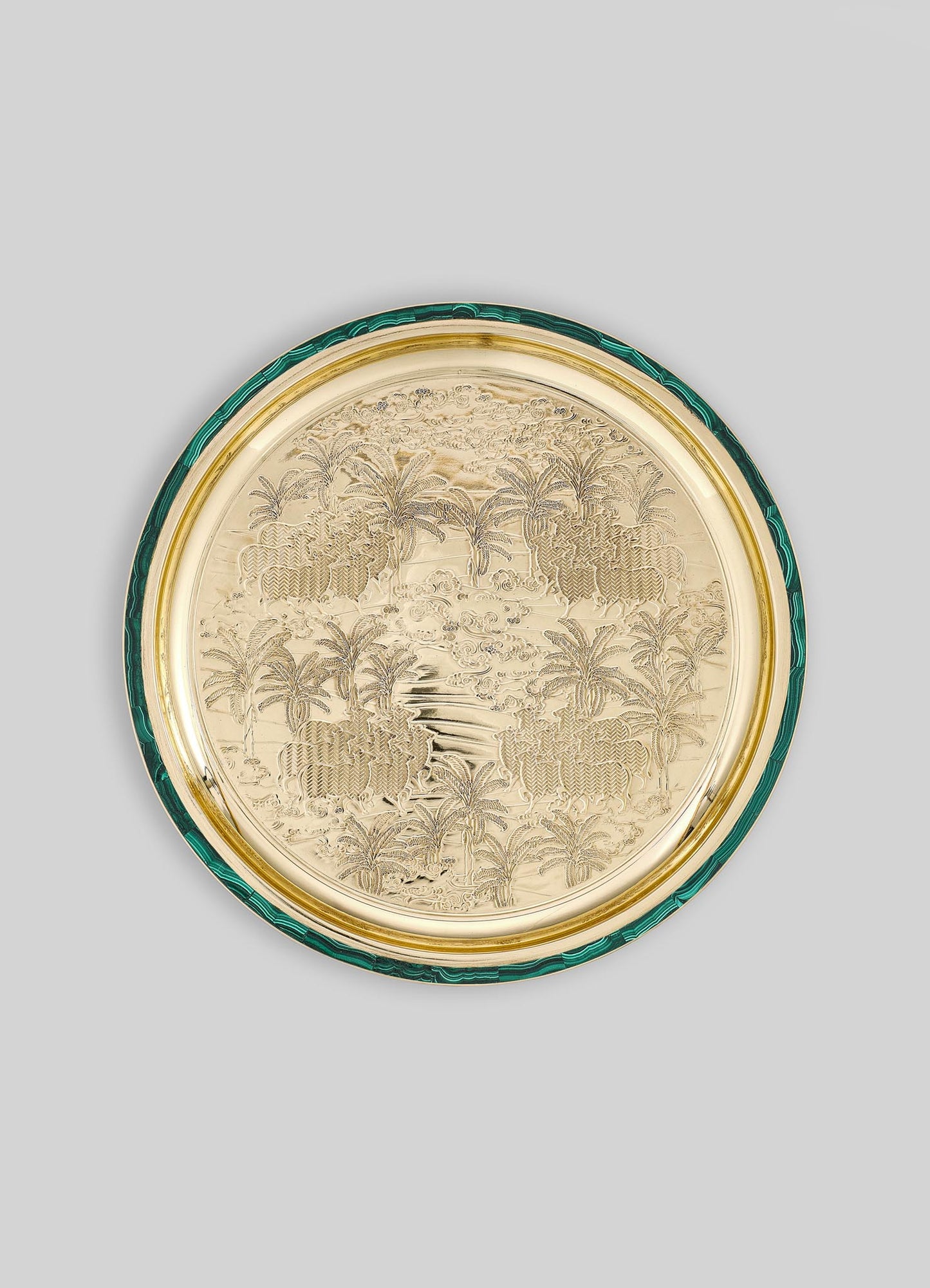A decorative round tray with an engraved tropical landscape featuring palm trees, a river, and animals, crafted in a metallic gold finish with a green malachite-patterned border.
