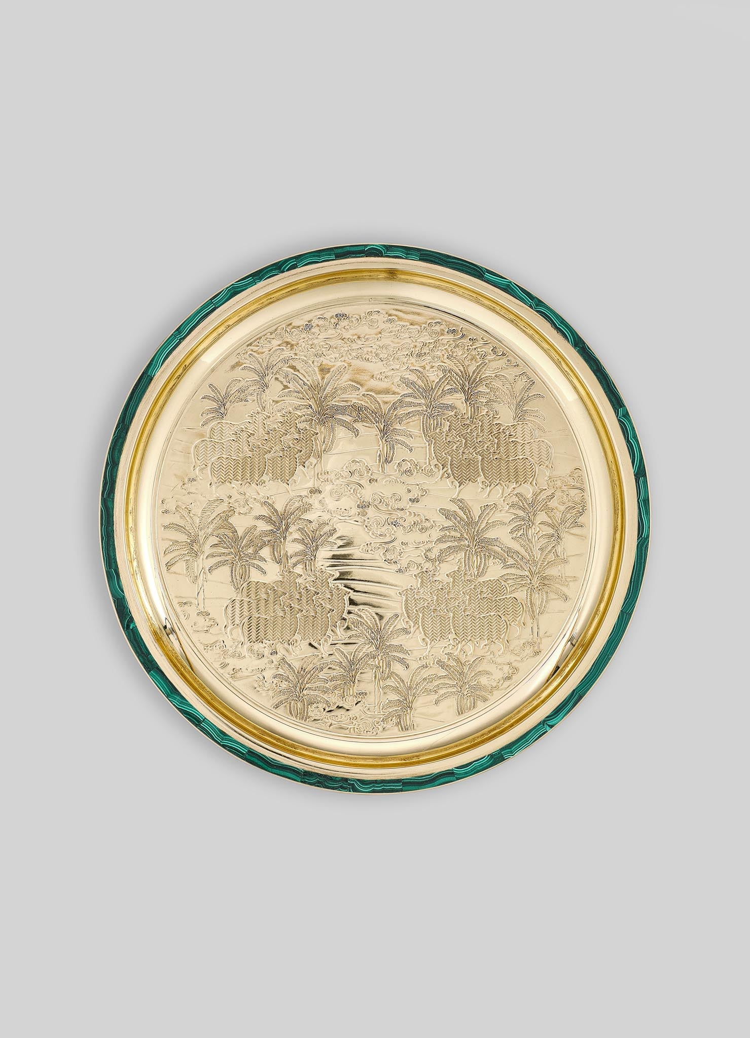 A decorative round tray with an engraved tropical landscape featuring palm trees, a river, and animals, crafted in a metallic gold finish with a green malachite-patterned border.
