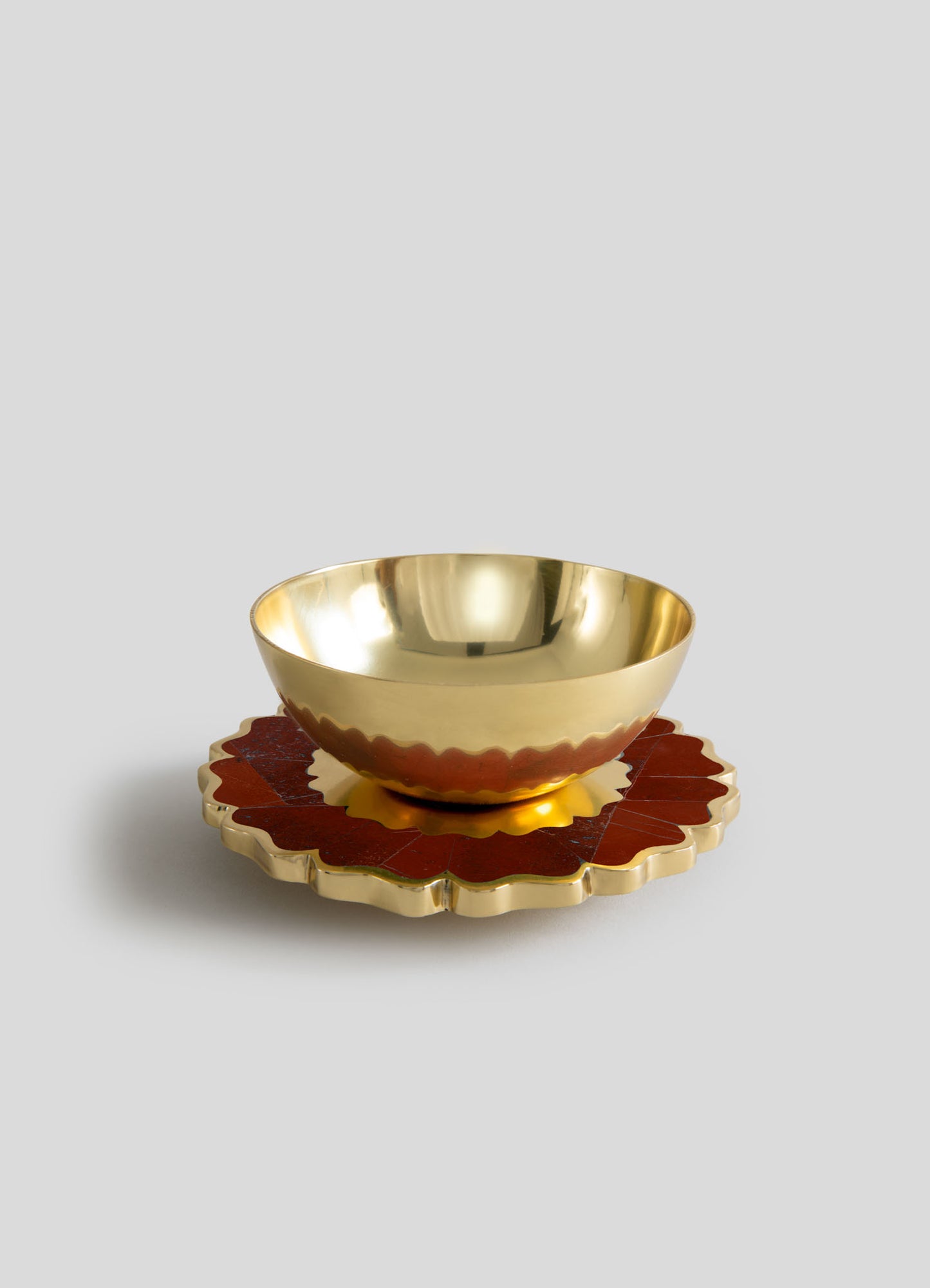 A polished brass bowl with a reflective surface, placed on a decorative scalloped-edged coaster with a red and gold pattern.