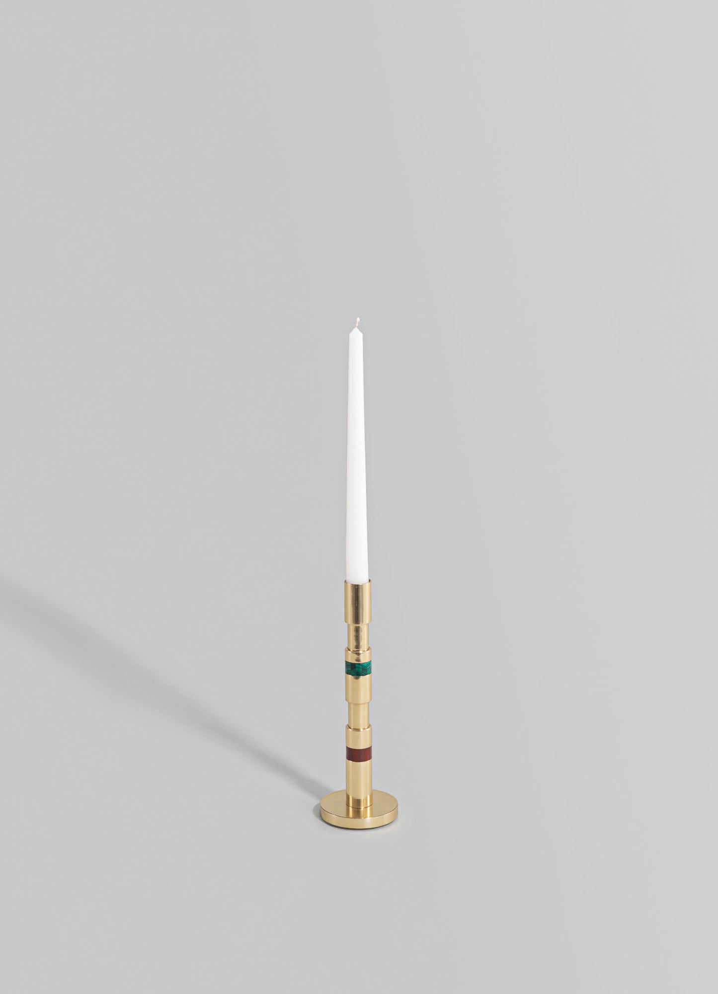A tall, slim candlestick holder made of metal with a brushed gold finish, featuring colorful geometric accents in green and burgundy, holding a white taper candle.