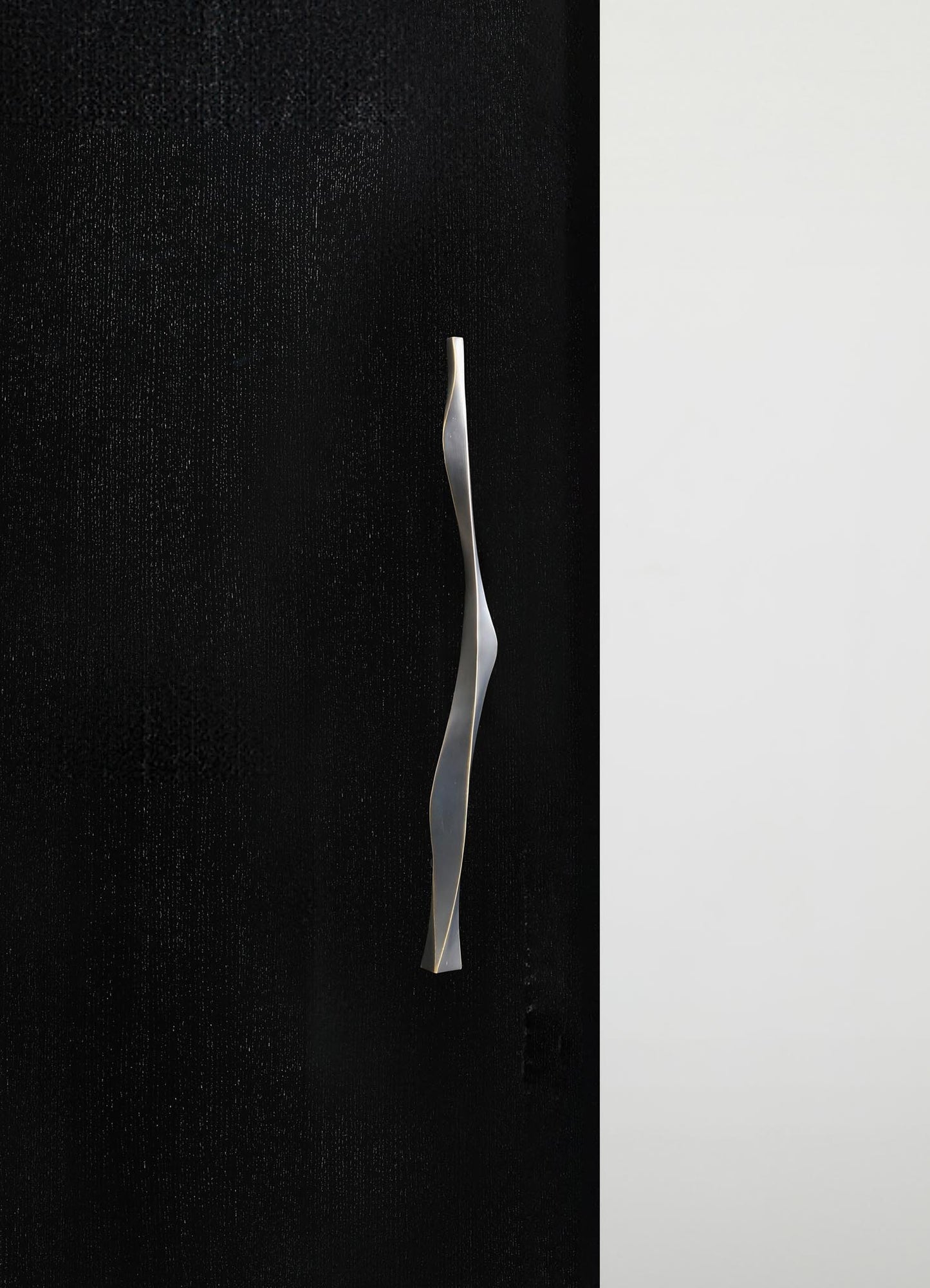 A sleek, modern door handle with a twisted, wavy design in matte metallic finish, mounted on a black textured door panel with a contrasting white wall background.