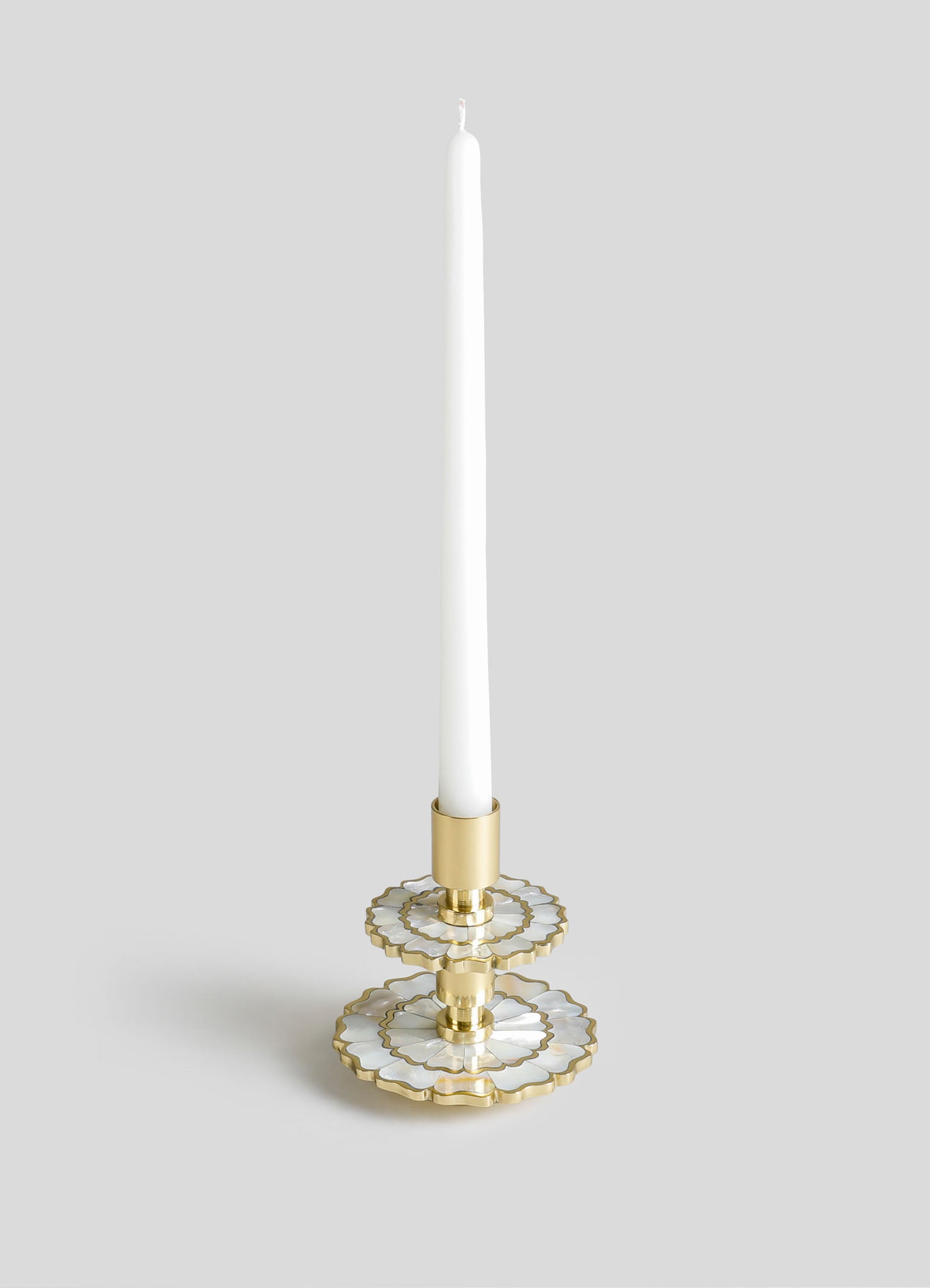 A tall, white taper candle mounted in a decorative candle holder with a layered, floral-inspired design in gold and white metal. The candle holder features scalloped edges and a polished finish, creating a modern and elegant aesthetic.