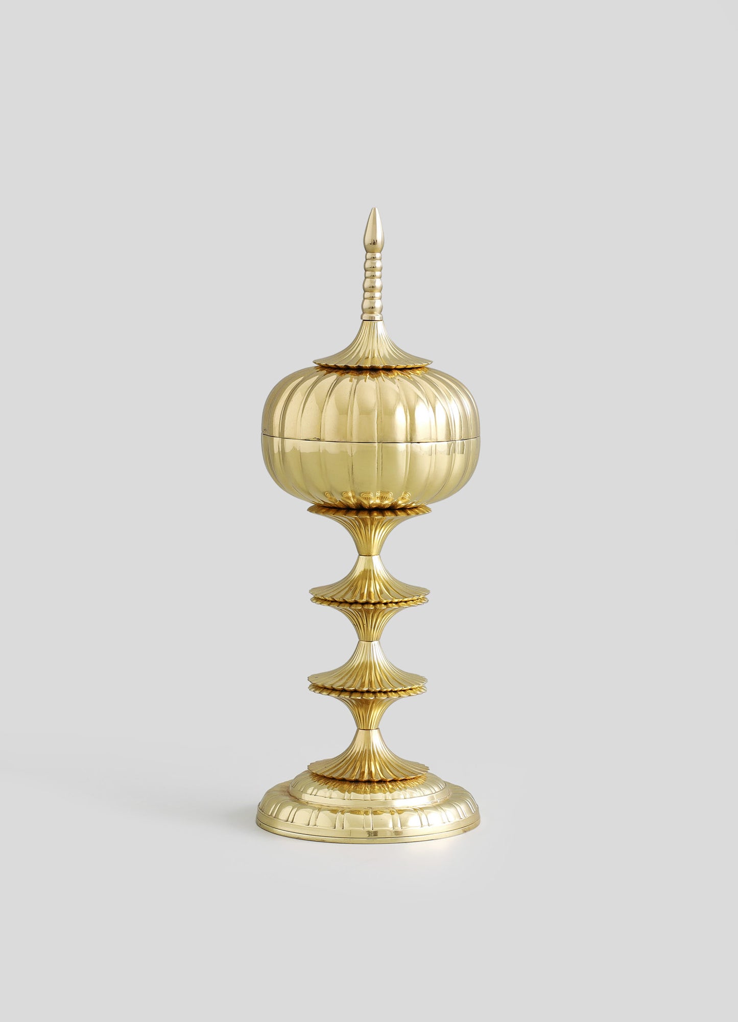 A decorative brass container with a rounded, fluted body and a pointed, ornate lid, mounted on a tall, tiered stand with scalloped edges. The piece features a polished gold finish, intricate ridges, and a classical style.