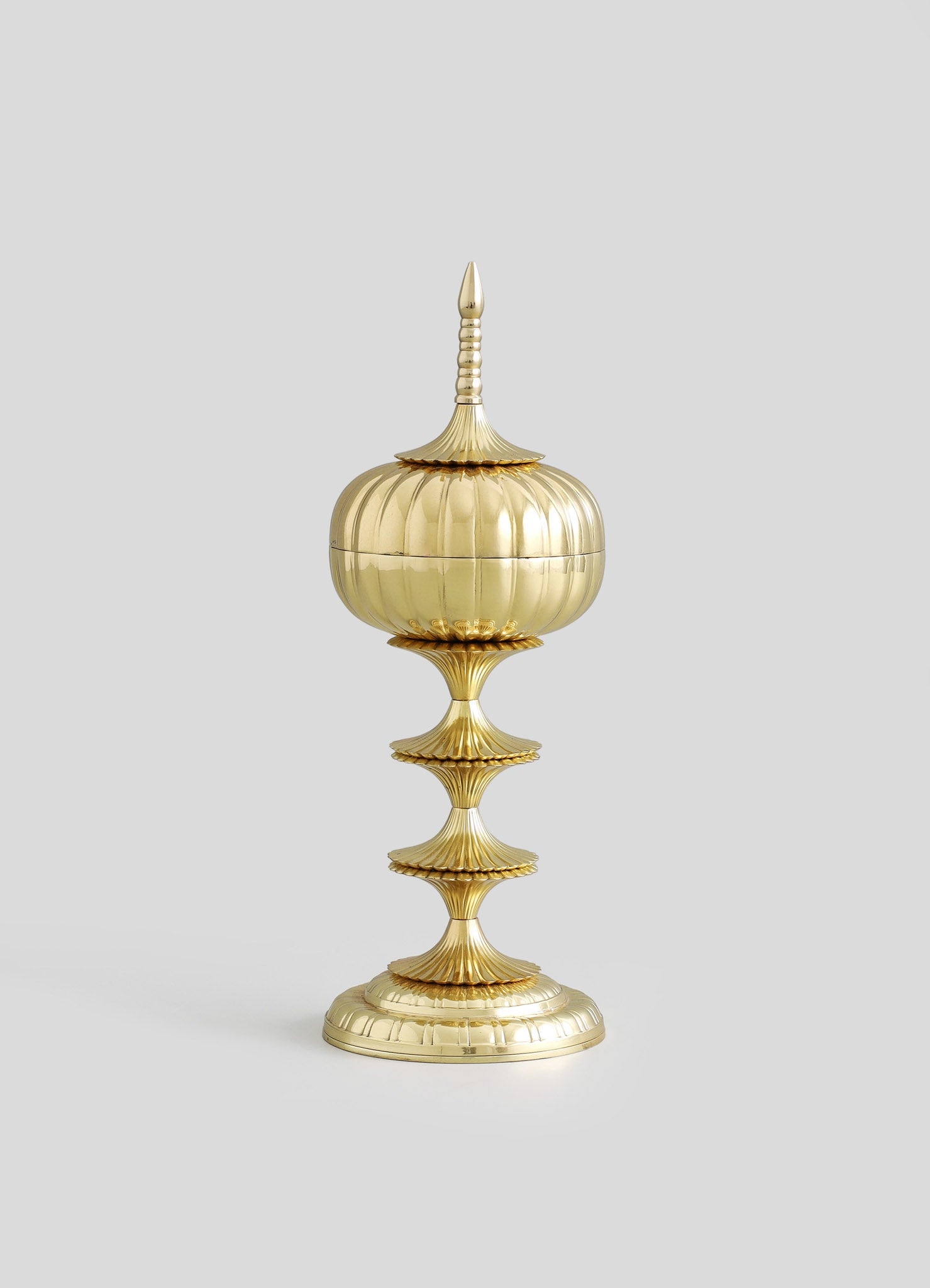 A decorative brass container with a rounded, fluted body and a pointed, ornate lid, mounted on a tall, tiered stand with scalloped edges. The piece features a polished gold finish, intricate ridges, and a classical style.