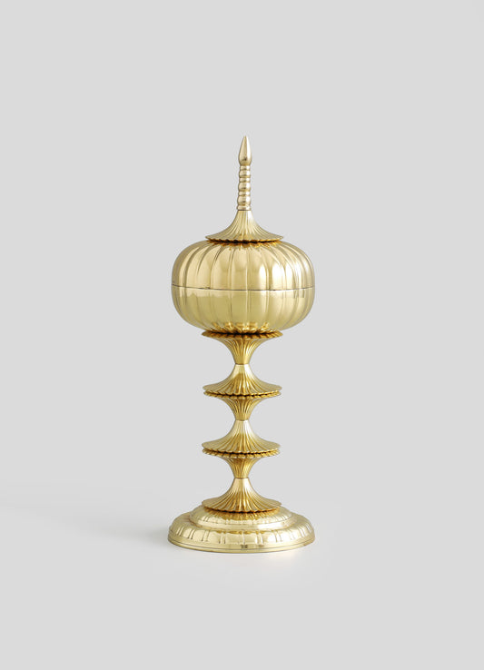 A decorative brass container with a rounded, fluted body and a pointed, ornate lid, mounted on a tall, tiered stand with scalloped edges. The piece features a polished gold finish, intricate ridges, and a classical style.