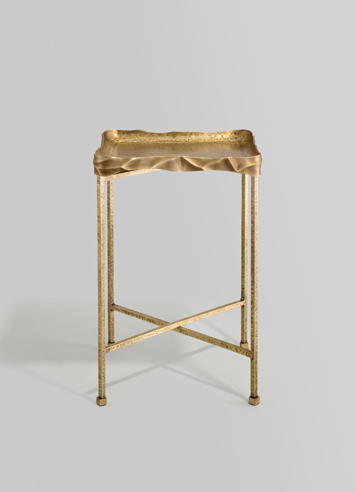 A gold-toned metal side table with irregular, wrinkled edges on the tabletop and three straight, slender legs, featuring a crossbar support. The style appears contemporary and sculptural.