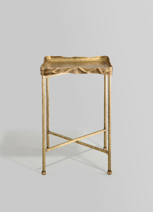 A gold-toned metal side table with irregular, wrinkled edges on the tabletop and three straight, slender legs, featuring a crossbar support. The style appears contemporary and sculptural.