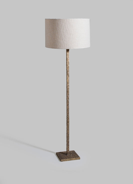 A floor lamp with a textured gold-tone metal base and stand, topped with a cylindrical beige linen lampshade.
