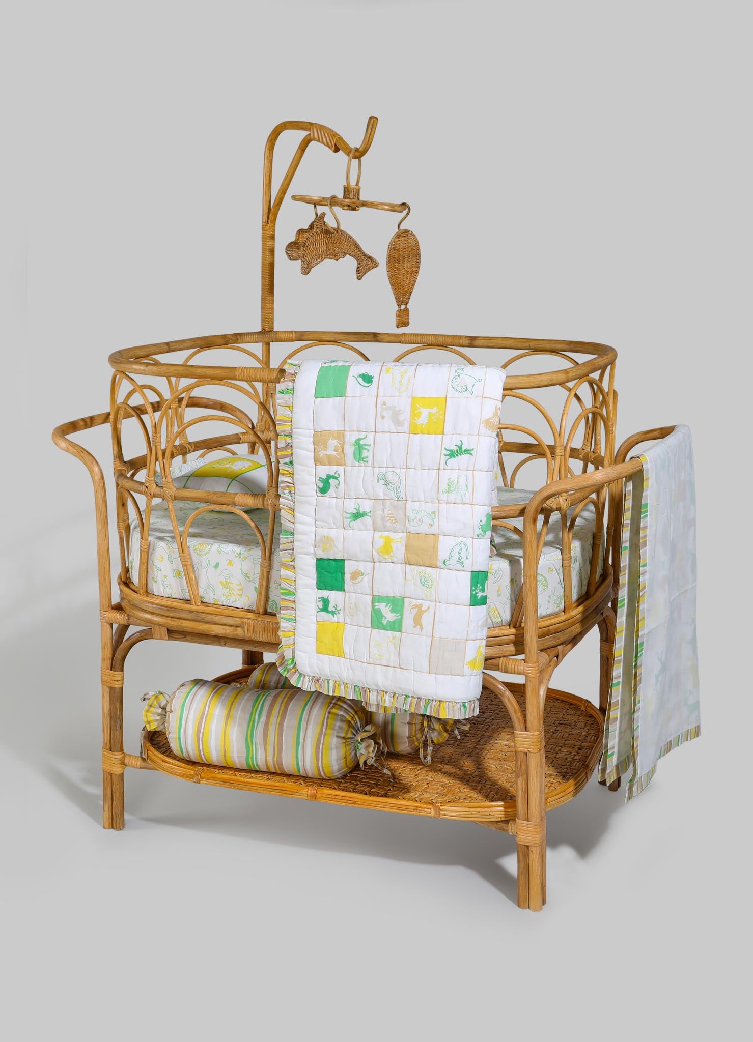 A woven rattan crib with a rounded frame and intricate detailing, featuring a mattress covered with patterned fabric. It includes hanging accessories shaped like aquatic animals and a fabric canopy. The bedding and accessories are decorated with colorful prints, enhancing a coastal or tropical style.