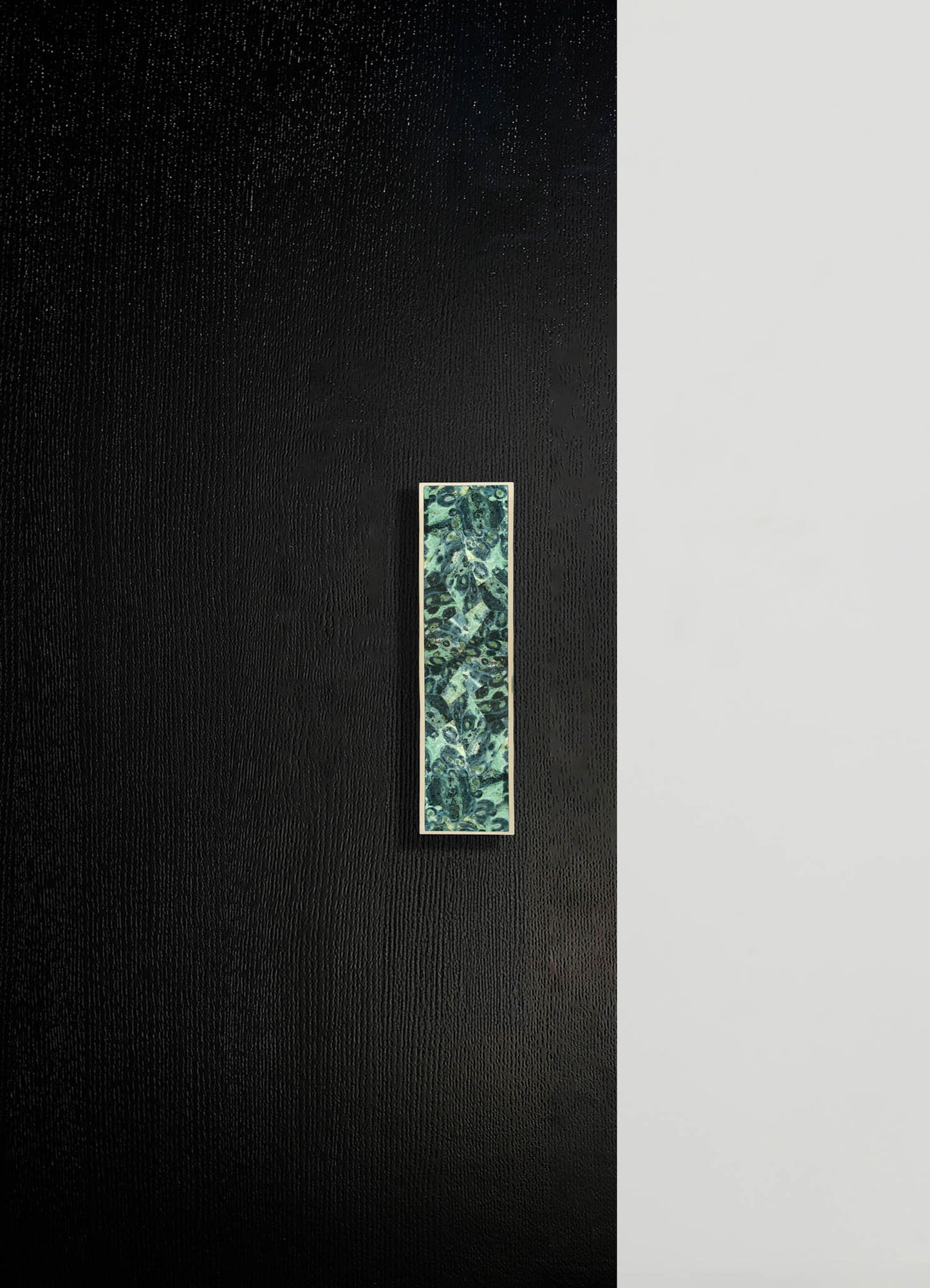 A minimalist interior wall with black textured and smooth light gray surfaces, featuring a rectangular decorative panel with a green and blue abstract pattern mounted vertically.