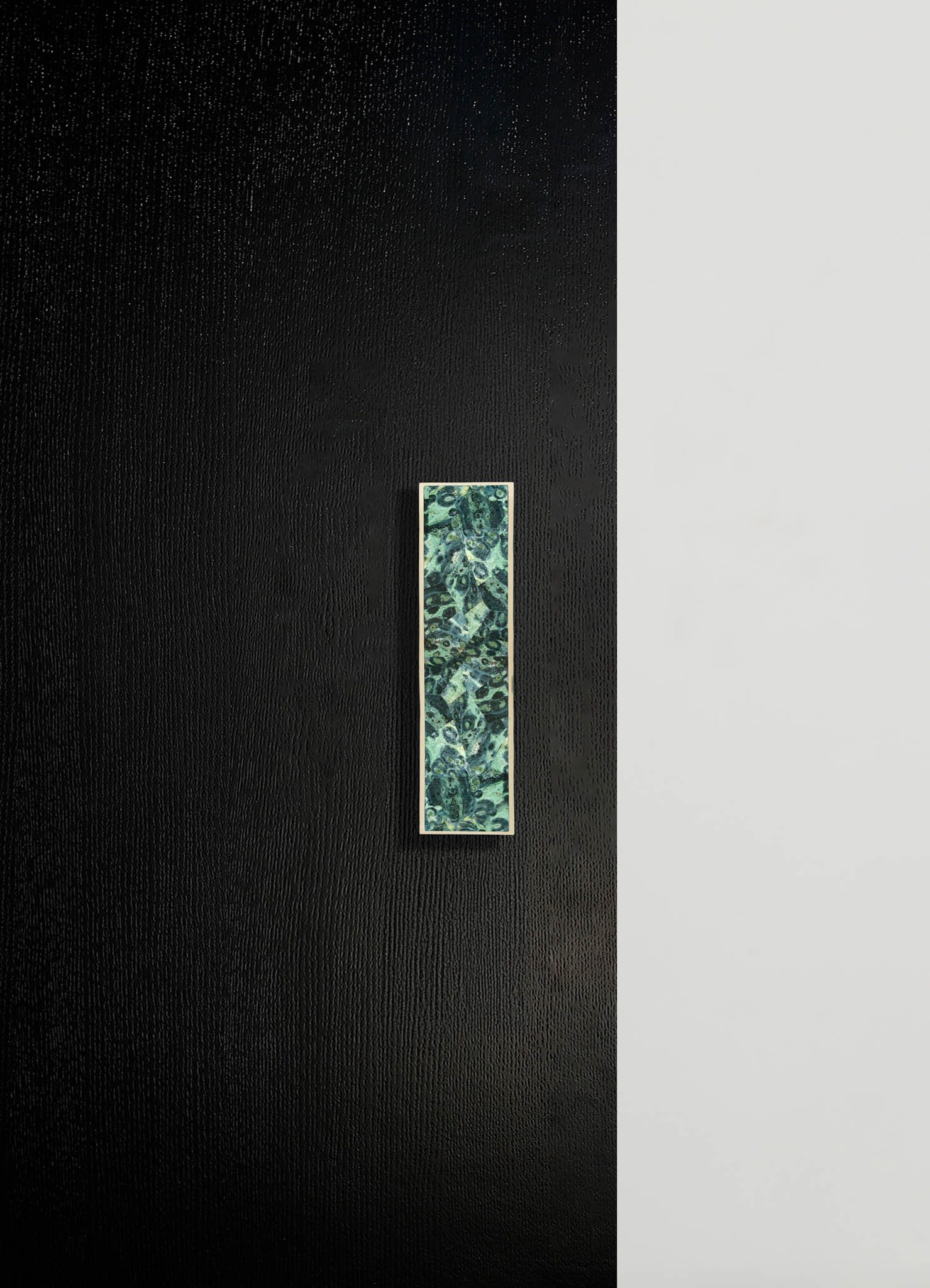 A minimalist interior wall with black textured and smooth light gray surfaces, featuring a rectangular decorative panel with a green and blue abstract pattern mounted vertically.