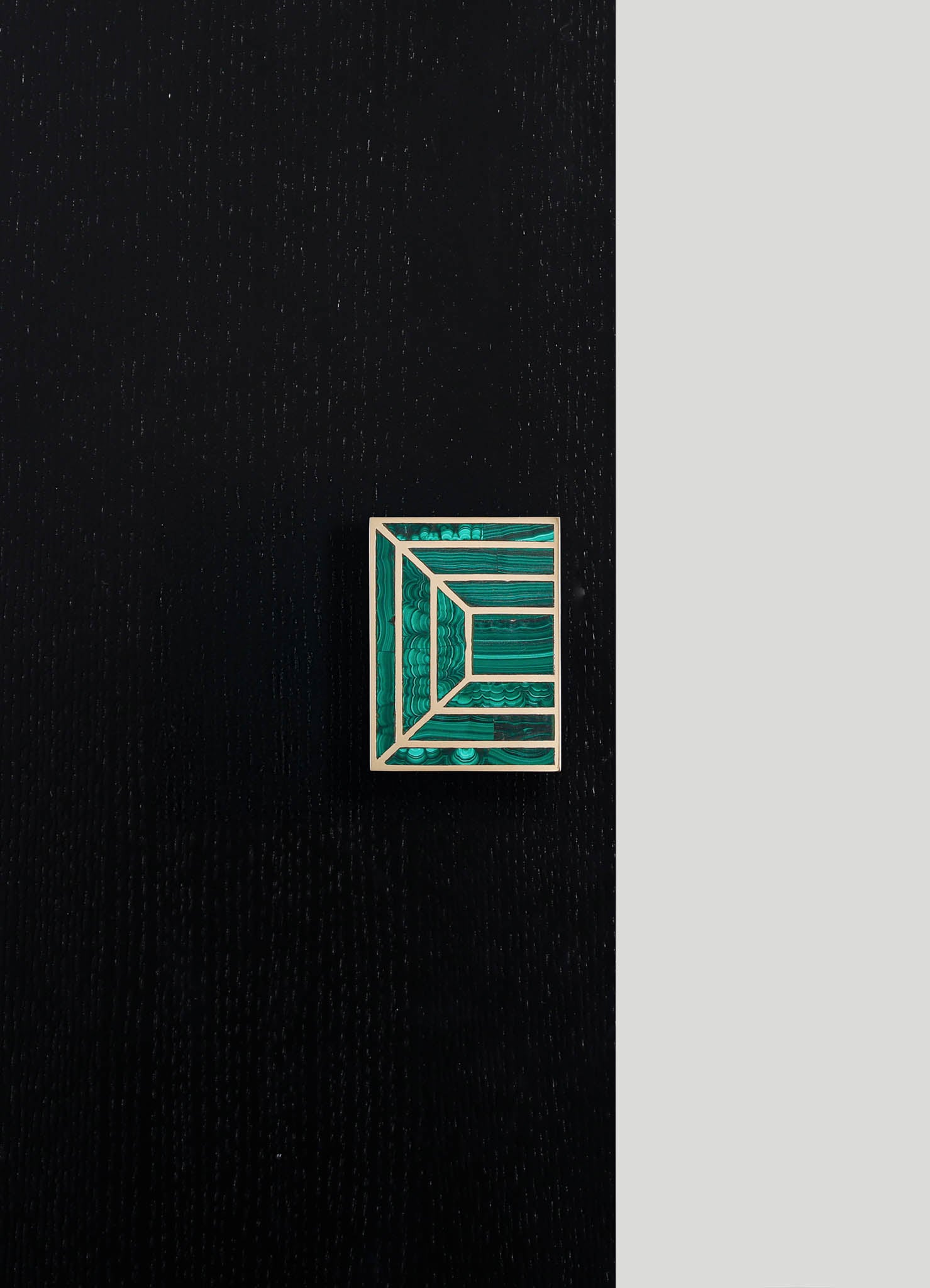 A rectangular jewelry box with a geometric design featuring crossed lines and rectangles in shades of green and blue, framed by a light-colored border, placed on a dark textured surface with a contrasting light background.