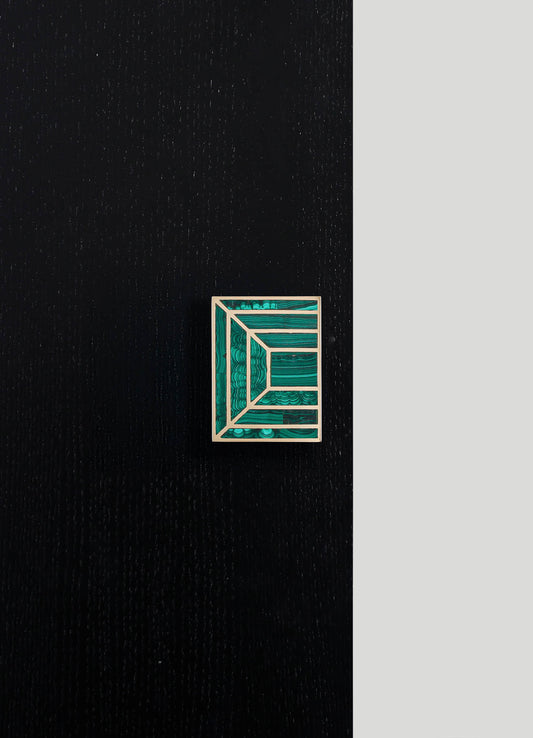 A rectangular jewelry box with a geometric design featuring crossed lines and rectangles in shades of green and blue, framed by a light-colored border, placed on a dark textured surface with a contrasting light background.