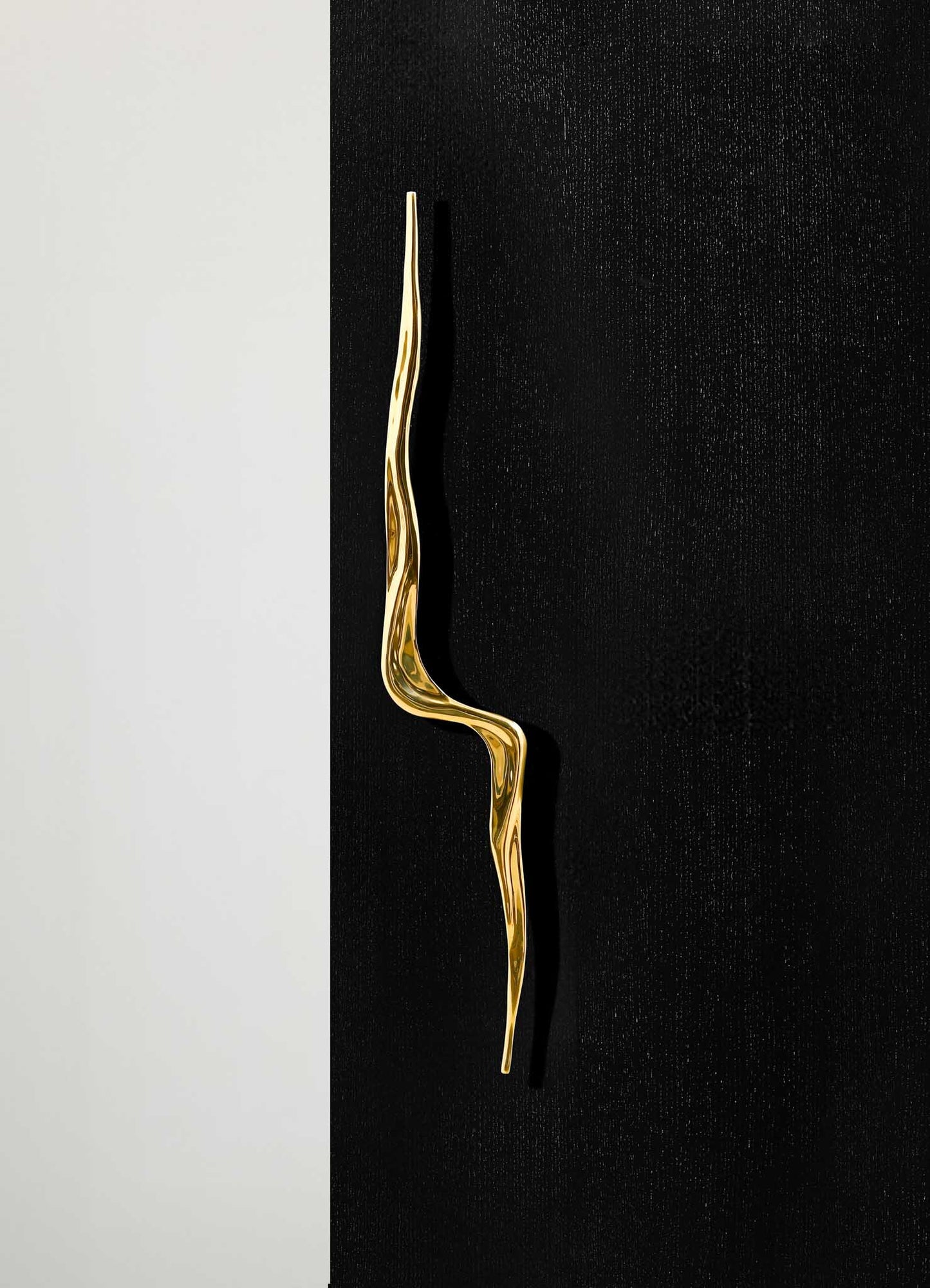 A gold-colored metal wall hook with an irregular, organic shape mounted on a black wall. The background features a narrow vertical strip of light-colored wall, creating a contrast with the textured black surface.