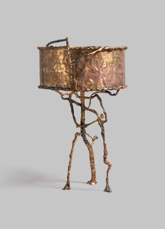 Decorative table lamp with a sculptural, organic metal base resembling twisted branches, finished in a textured, metallic rose gold tone. The cylindrical lampshade appears to be made of hammered metal with a similar rose gold finish, creating a rustic yet contemporary aesthetic.