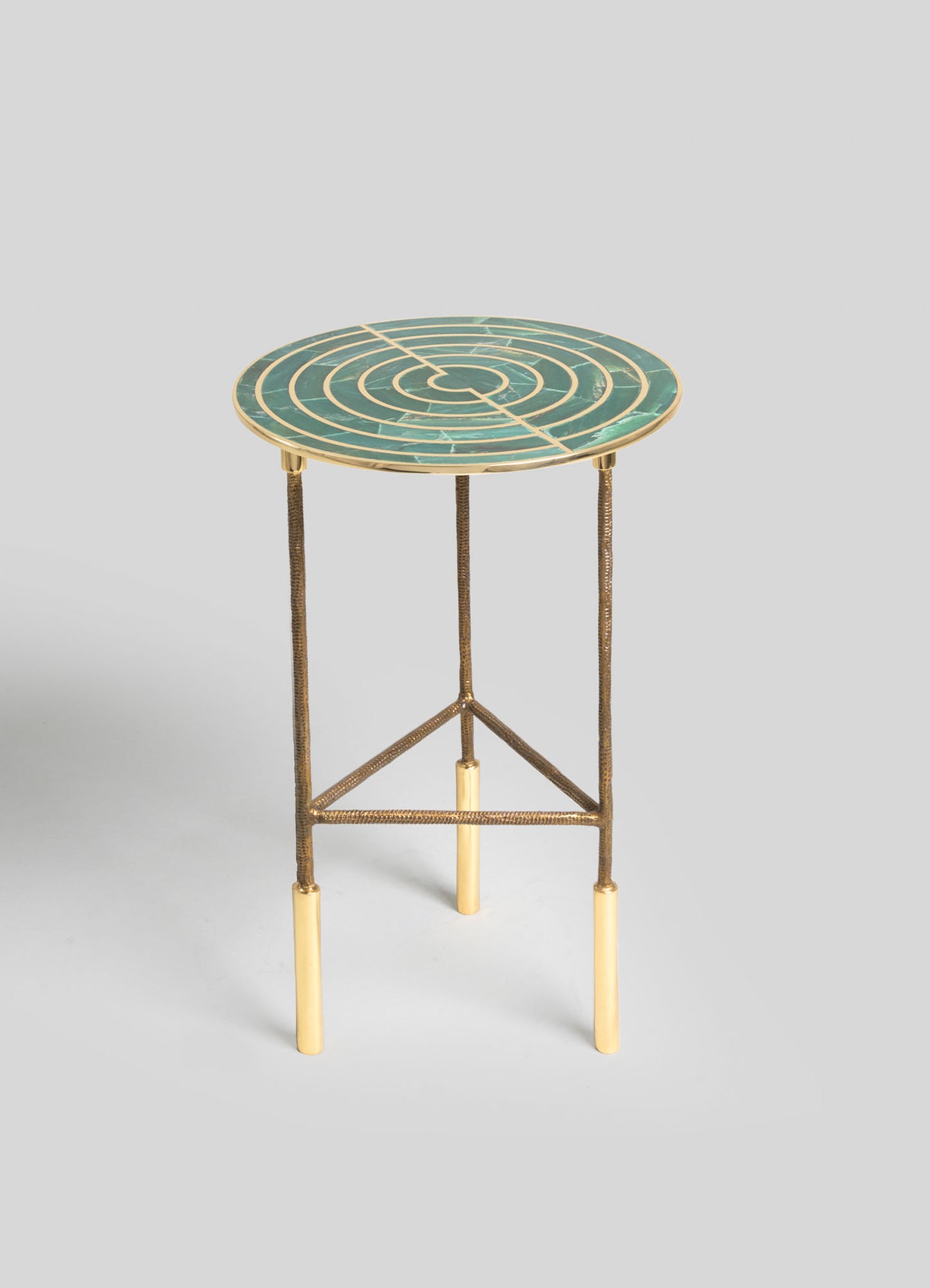 A round side table with a green marble top featuring concentric circles pattern and brass trim, supported by slender metal legs with a textured finish and light-colored bases, all set against a plain background.