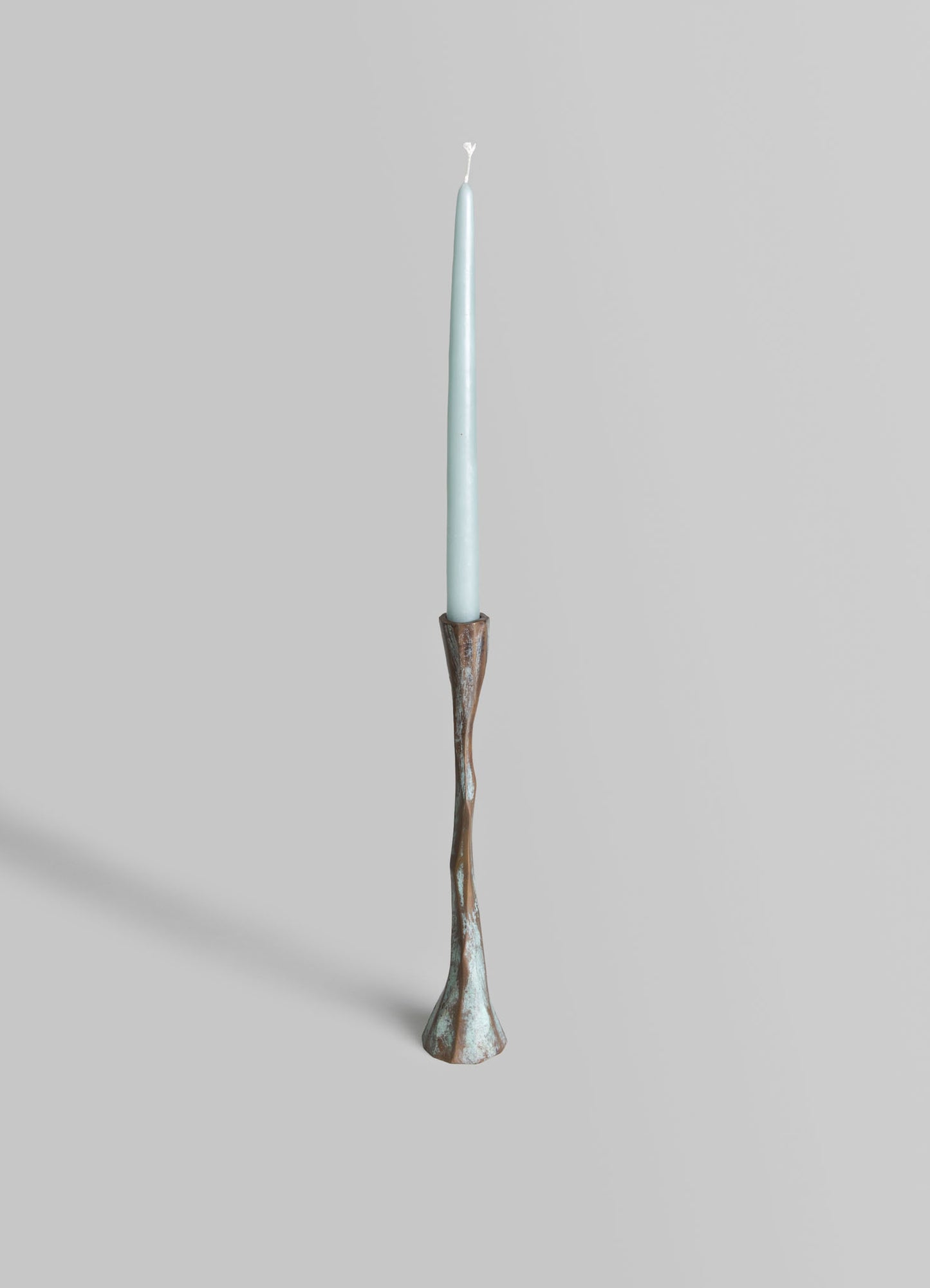 A tall, slender candle holder with a carved, organic shape in distressed brown and turquoise finish, holding a light blue taper candle.