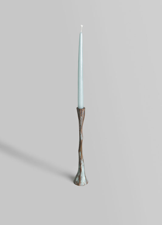 A tall, slender candle holder with a carved, organic shape in distressed brown and turquoise finish, holding a light blue taper candle.