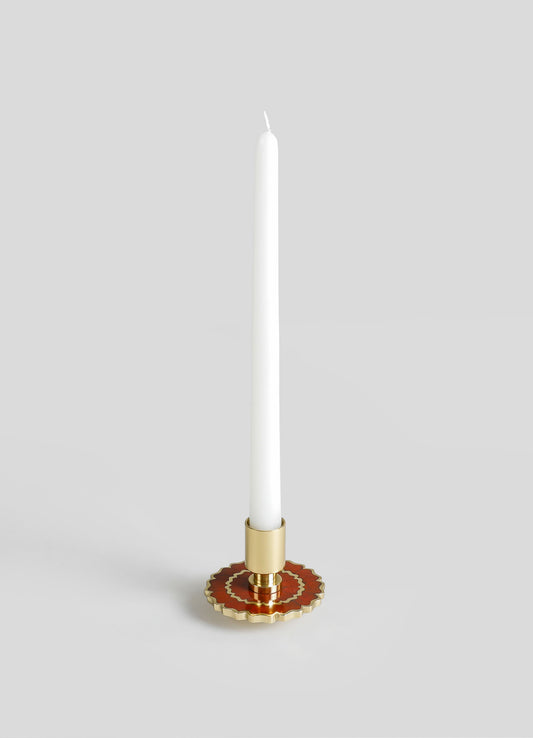 A tall white taper candle in a decorative candle holder with a scalloped edge, featuring gold and reddish-brown accents, set against a plain gray background.