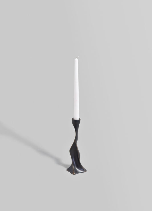 A tall white taper candle in a sculptural black metal candle holder with an organic, flowing design, set against a neutral gray background.