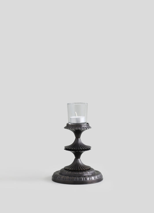 A black metal candle holder with a ribbed, tiered design and a round base, supporting a glass container with a white pillar candle inside.