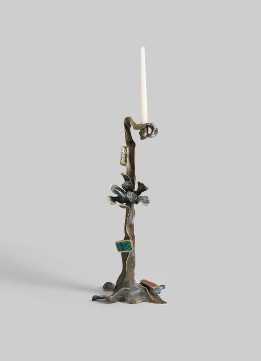 A sculptural candle holder made of metal with an organic, branch-like design, featuring a single white taper candle at the top. The piece has a bronze finish with textured, leaf-like details and small colorful accent elements.