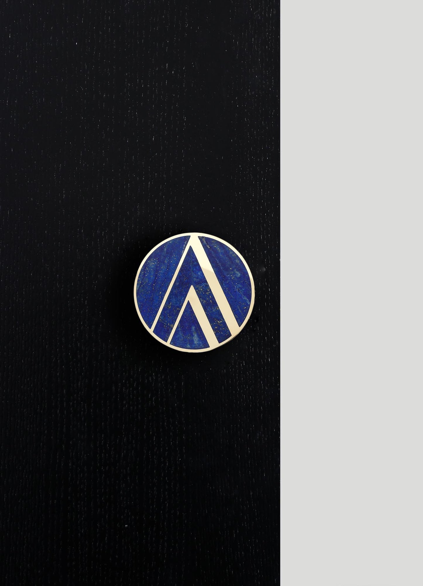 A round enamel pin with a geometric design in white and shades of blue, placed on a dark textured fabric background.