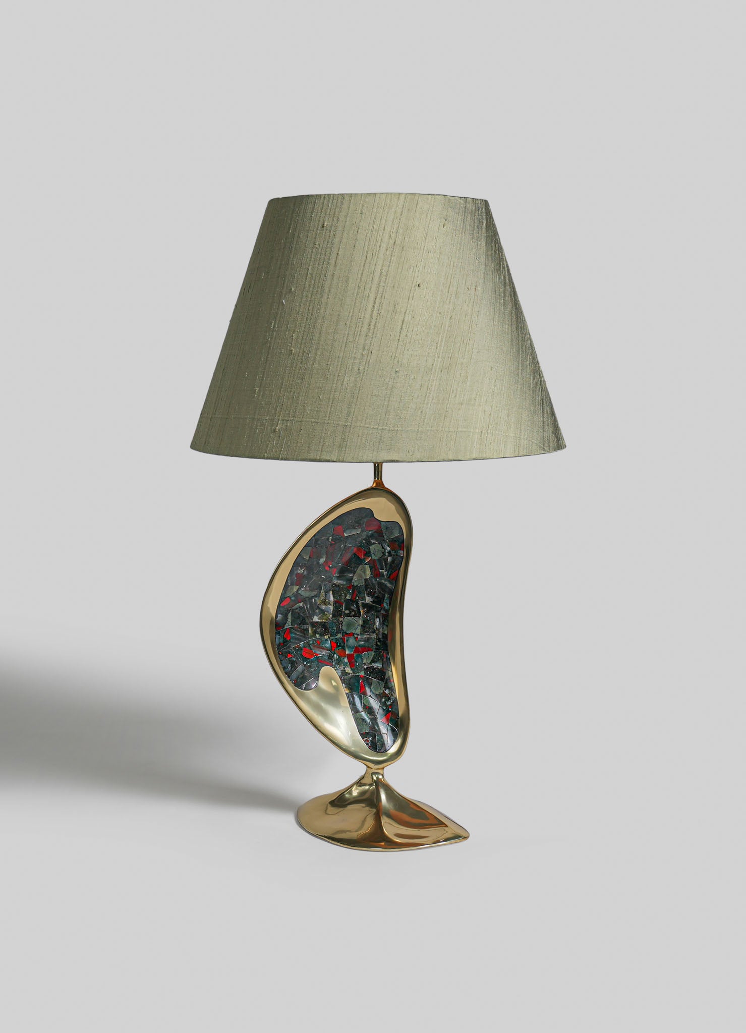 A table lamp with a beige fabric lampshade and a sculptural base featuring a mixed-material design in gold and mosaic glass fragments, set against a plain background.