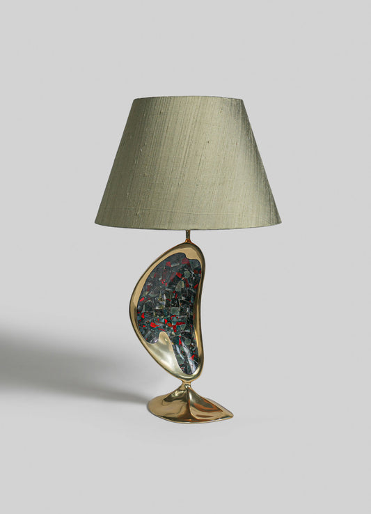 A table lamp with a beige fabric lampshade and a sculptural base featuring a mixed-material design in gold and mosaic glass fragments, set against a plain background.